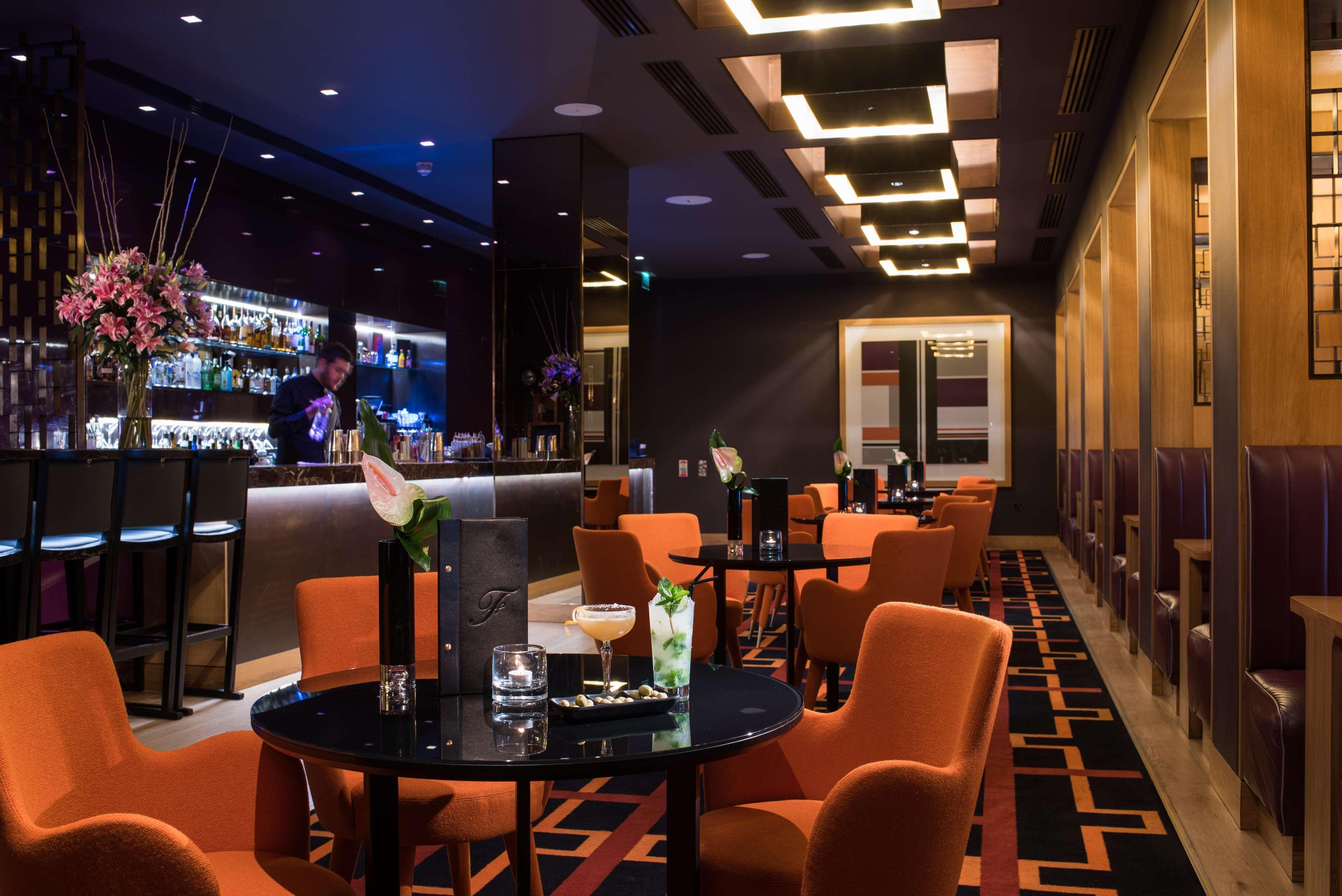 ザ フィッツウィリアム ホテル ベルファスト エクステリア 写真 The photo depicts a stylish bar or lounge area with a modern and sophisticated atmosphere. The space features dark walls accented by blue lighting and a sleek bar counter in the background, stocked with various drinks. The seating consists of round t