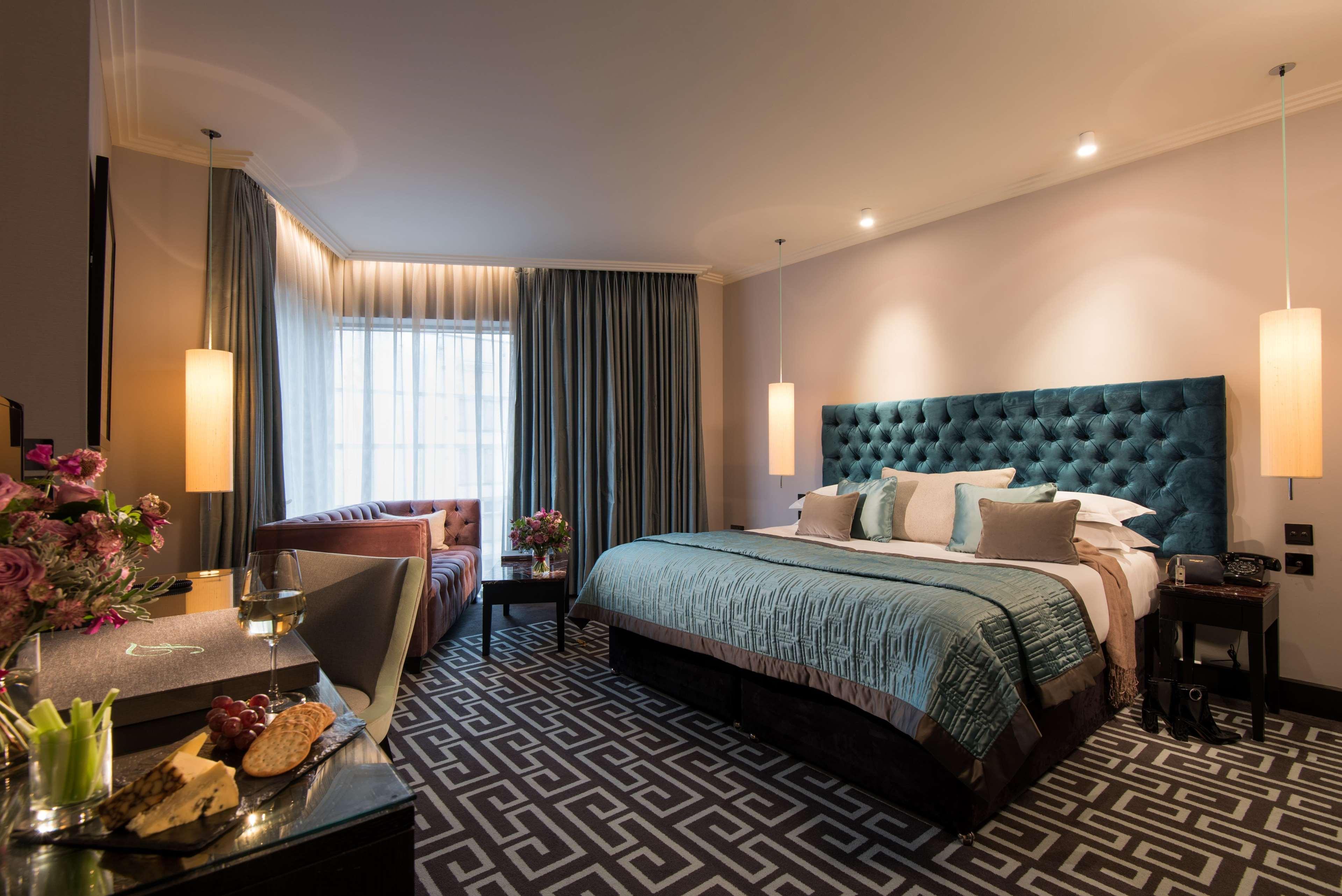 ザ フィッツウィリアム ホテル ベルファスト エクステリア 写真 The photo depicts a stylish hotel room. It features a large bed with a tufted teal headboard, flanked by two wall sconces providing soft lighting. The bedding is neatly arranged with accent pillows. To one side, there is a small seating area with a c