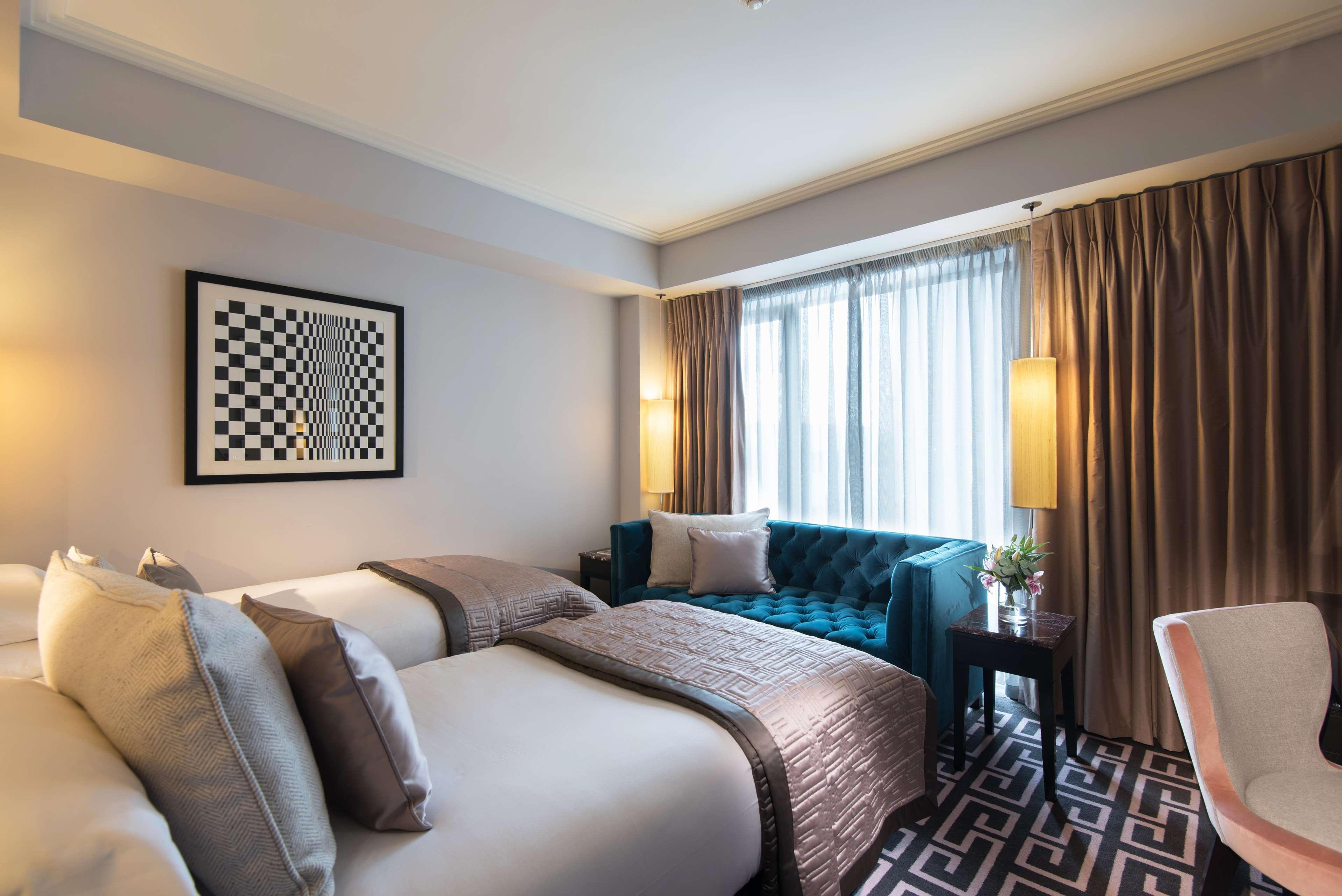 ザ フィッツウィリアム ホテル ベルファスト エクステリア 写真 The photo shows a modern hotel room. It features two beds with decorative pillows, a stylish teal couch, and large windows with sheer curtains allowing natural light to enter. The walls are painted in neutral tones, and there is a piece of artwork wi