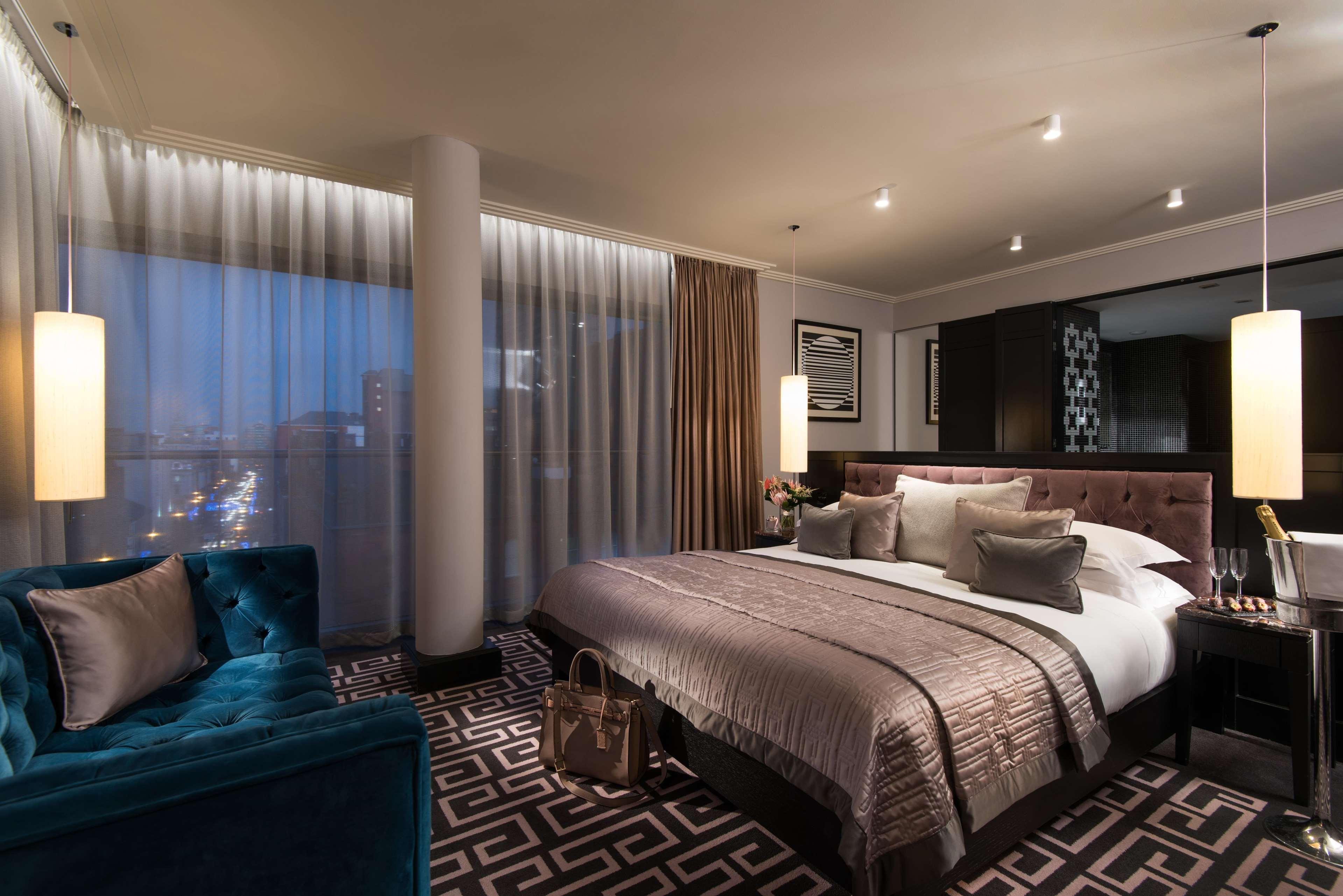 ザ フィッツウィリアム ホテル ベルファスト エクステリア 写真 The photo shows a luxurious hotel room with a modern design. It features a large bed with decorative pillows and a stylish bedspread, complemented by bedside lamps. To one side, there is a plush turquoise sofa and a coffee table. The room has floor-t