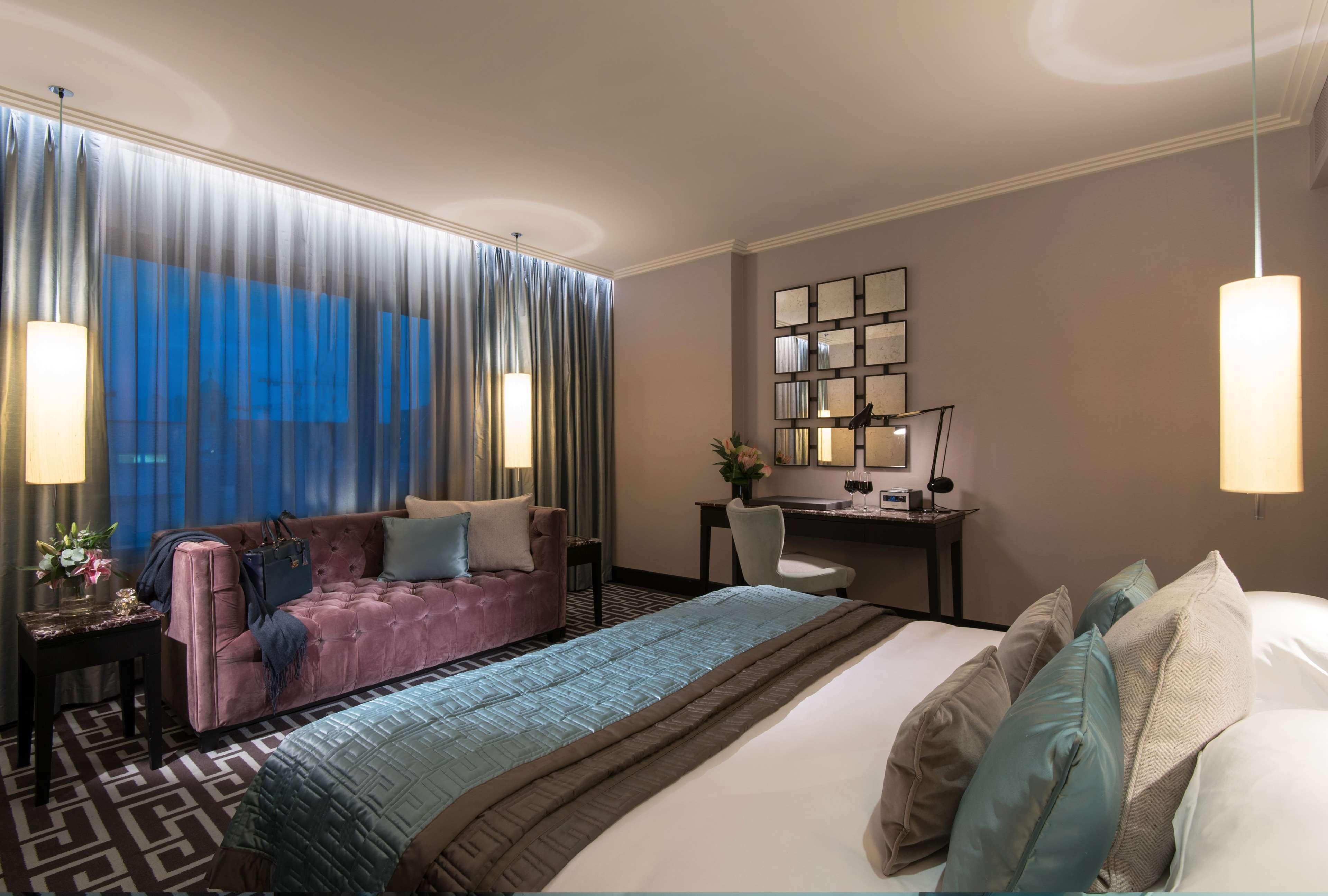 ザ フィッツウィリアム ホテル ベルファスト エクステリア 写真 The photo depicts a modern hotel room featuring a spacious layout. There is a large bed with decorative pillows in the foreground, covered with a stylish bedspread. To one side, a plush purple sofa adds a pop of color and comfort. A small desk and ch